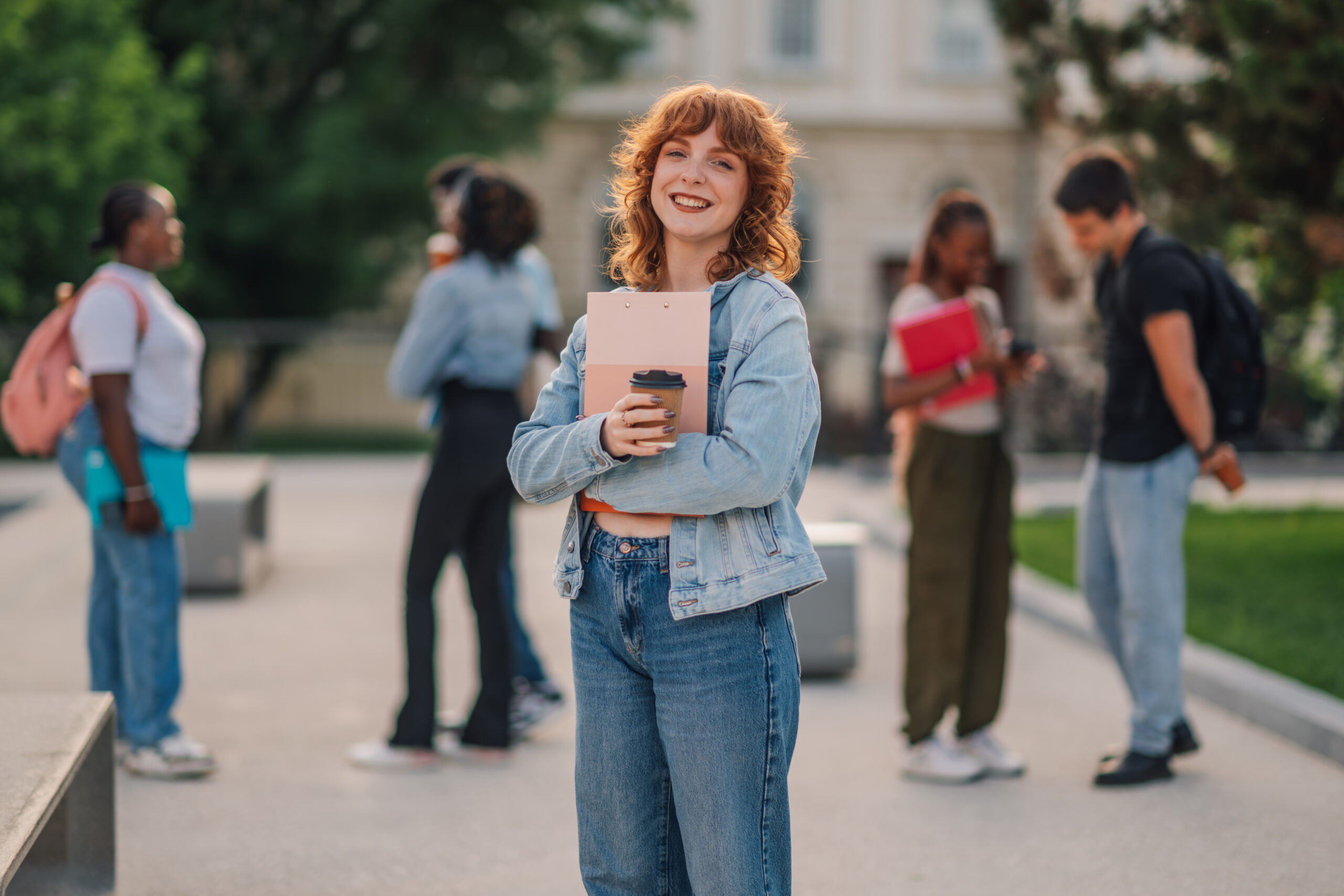 Three quarter length portrait of adolescent ginger college girl standing at university campus with textbook and takeaway coffee in hands and smiling at the camera while having coffee break at academy.